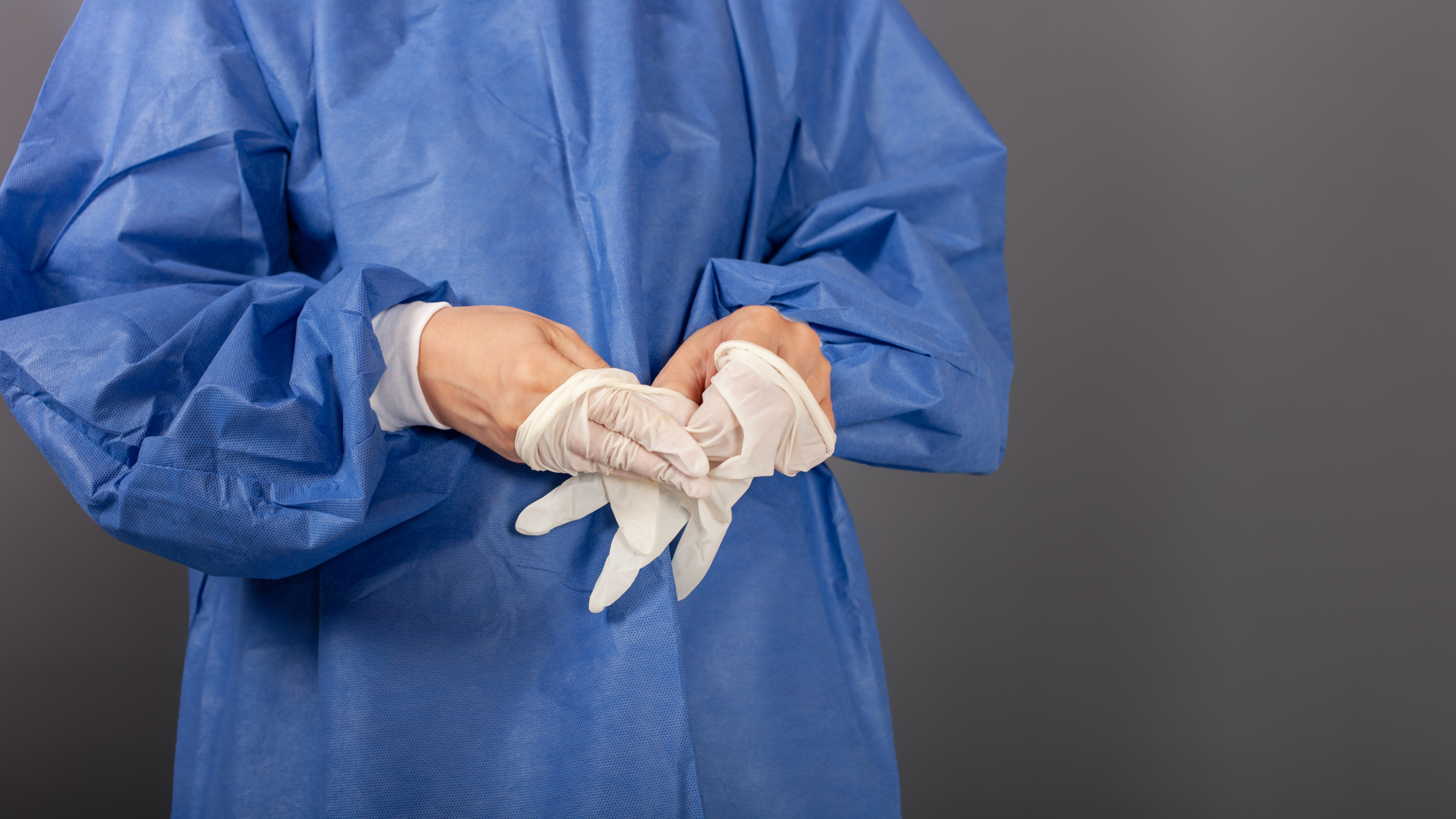 A woman doctor takes off her white gloves. A woman in a blue surgical robe, a gray background, an empty place